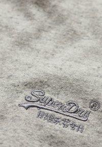 Grå sweatshirtstof med en tekstureret overflade, udstyret med et broderet "SuperDry" logo i mørkegrå. Nærbillede af logodetaljer synligt.