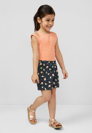 Jeune fille souriante aux cheveux foncés attachés en queue de cheval, portant un haut sans manches pêche, un short bleu marine à motifs et des sandales métalliques, marchant à l'intérieur.