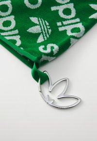 Zaļš kokvilnas dvielis ar baltu "adidas" tekstu un logo; ietver sudraba metāla akcentu, kas ir veidots kā zīmola logo, un zaļu auduma cilpu.