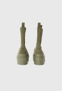 Olivengrønne ruskinds ankelstøvler med trækflapper, tykke gummisåler og en struktureret finish med "Converse All Star" branding på hælen.