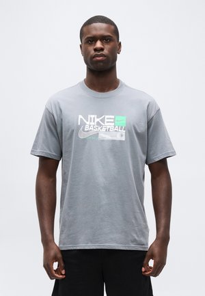 Νέος άνδρας που στέκεται φορώντας γκρι μπλουζάκι μπάσκετ Nike με λευκό και πράσινο λογότυπο, μπροστά σε απλό φωτεινό φόντο.