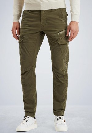 Homme portant un pantalon cargo vert olive avec des poches latérales et des baskets blanches, sur un fond uni.