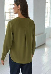 Blouse verte olive à manches longues avec un ourlet incurvé et un détail de pli au dos. Accompagnée d'un pantalon en denim foncé. Texture lisse.