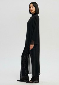 Sort langærmet kjole med høj hals, kombineret med skinnende sorte paliet-bukser med vide ben og sorte spidse sko. Glat tekstur.