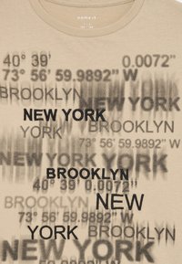 Beige T-Shirt mit Grafikdruck, der "NEW YORK" und "BROOKLYN" in Schwarz sowie geografische Koordinaten in unterschiedlicher Opazität zeigt.