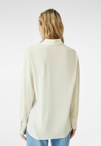 Kremowa, długorękawowa koszula o fakturowanym wykończeniu, z klasycznym kołnierzykiem i zapięciem na guziki, w zestawie z jasnoniebieskimi dżinsami.