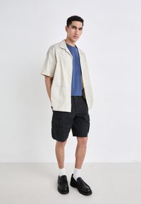 Camisa de manga corta color beige claro, camiseta azul debajo, pantalones cortos cargo negros, mocasines negros, calcetines blancos; atuendo casual con bolsillos y detalles de botones.