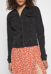 Kvinna som bär en svart denimjacka över en orange blommig kjol med en hög slits vid låret, visad från halva ansiktet till mitten av låret.