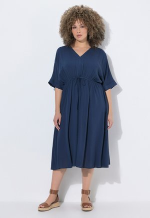 Frau mit lockigem Haar trägt ein marineblaues Midikleid mit tailliertem Schnitt und braune Keilsandalen mit Knöchelriemen und steht vor einer weißen Wand.