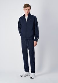 Navy tracksuit made of smooth fabric, featuring a zip-up jacket and elasticated pants, with logo accents on sleeves and sides.