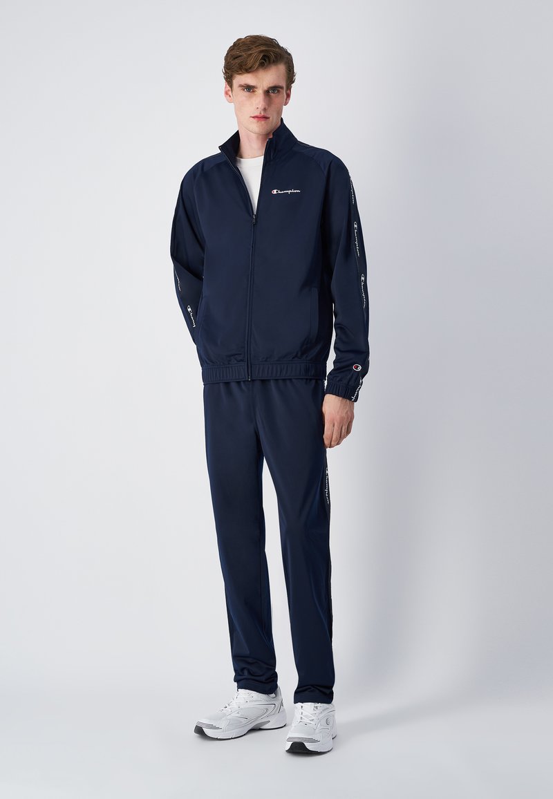 Chándal de color azul marino hecho de tela suave, que incluye una chaqueta con cremallera y pantalones elásticos, con acentos de logo en las mangas y los lados.