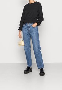 Schwarze Bluse mit langen Ärmeln und gerafftem Bund, kombiniert mit hellblauen gerade geschnittenen Jeans. Akzentuiert mit einer kleinen gelben Handtasche und schwarzen klobigen Schuhen.