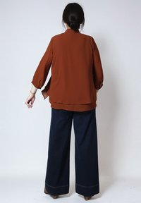 Vrouw met donker haar in een knot, gekleed in een roestkleurige blouse met lange mouwen en donkere wijde broek, staande met haar rug naar de camera op een witte achtergrond.