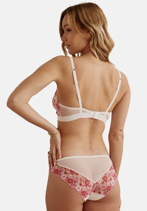 Femme portant une lingerie blanche transparente avec une broderie florale rose et rouge, vue de dos sur un fond uni.