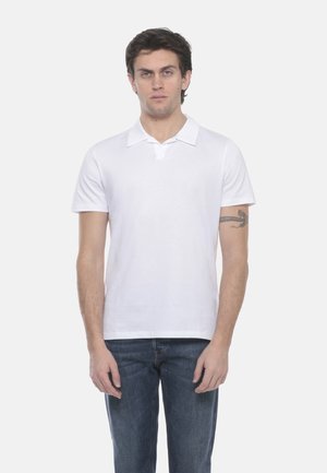 Giovane uomo con capelli scuri che indossa una polo bianca a maniche corte e jeans blu, in posizione frontale contro uno sfondo bianco.