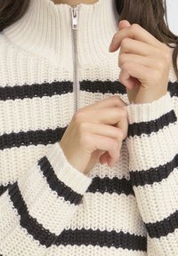 Pletený krémový sveter s čiernymi horizontálnymi pruhmi, vysokým golierom a strieborným zipsom. Žebrované manžety a štruktúrovaná textúra.