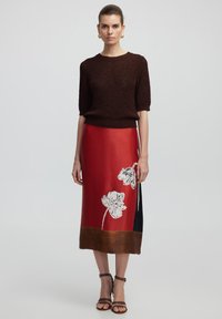Suéter de punto marrón, falda de satén roja con estampados de flores blancas y acentos marrones. Sandalias con correas en el tobillo completan el look.