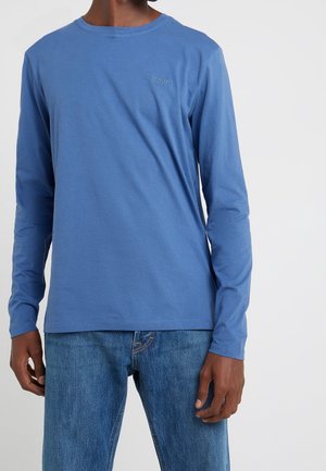 Homme portant une chemise bleue à manches longues avec un petit logo de marque sur la poitrine et un jean denim bleu moyen, sur un fond uni.
