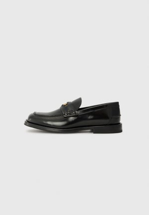 Mocassin en cuir noir avec un bout arrondi, des coutures décoratives et un petit rabat à l'avant. Semelle en caoutchouc plate avec un talon minimal.