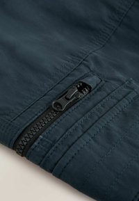 Gros plan sur un tissu bleu foncé avec une fermeture éclair noire et une couture détaillée, mettant en valeur la tirette de la fermeture éclair et la construction de la couture.