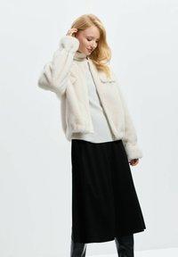 Biała sztuczna futrzana kurtka z zamkiem błyskawicznym, żeberkowym kołnierzem i szeroką czarną spódnicą. Gładka tekstura, luźny krój i lekko bufiaste rękawy.
