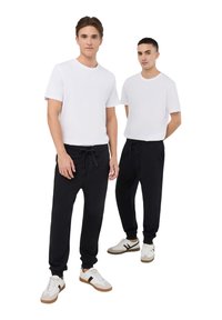 Uomini in t-shirt bianche a girocollo e pantaloni jogger neri, dotati di vita con cordoncino, tasche e polsini elasticizzati, abbinati a sneaker bianche.