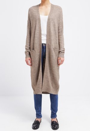 Femme portant un long cardigan beige ouvert devant, top blanc, jean bleu et mocassins noirs, debout sur un fond blanc.