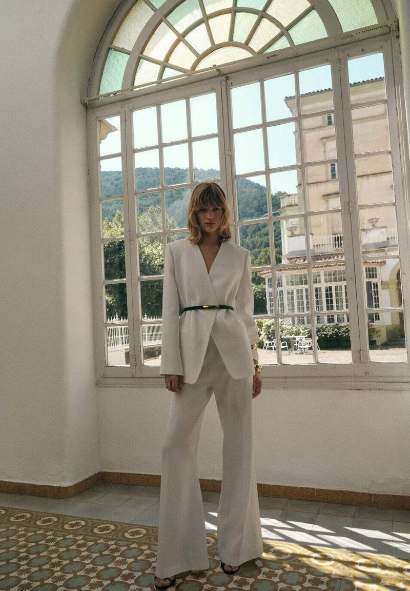 Witte maatpak met een getailleerde blazer, wijde broek en een dunne zwarte riem. Glad materiaal met een lichte glans en minimalistisch ontwerp.