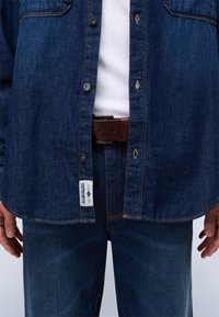 Camisa de mezclilla azul oscuro con frente abotonado y bolsillos en el pecho, combinada con jeans de mezclilla azul oscuro a juego. Cinturón de cuero marrón con hebilla visible.