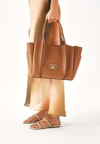 Borsa a tracolla in pelle marrone con finitura testurizzata, chiusura con cerniera e hardware dorato. Abbinata a sandali beige con cinturini e una gonna fluente in ombre.