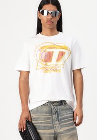 Hvit t-skjorte med grafisk trykk i gult og rødt, kombinert med lyse, slitte jeans og elegante svarte solbriller. Holder en svart veske.