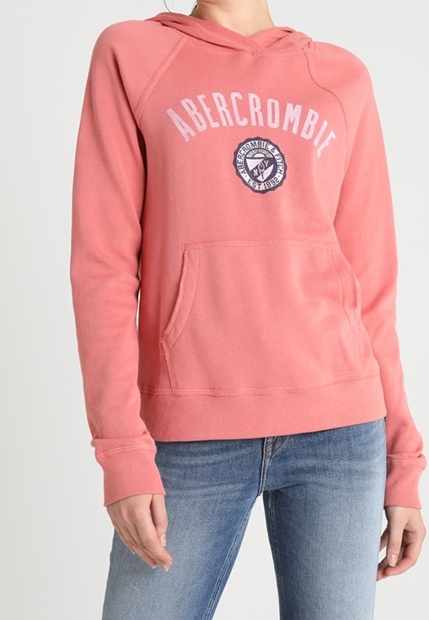 Žena v korálově růžové mikině Abercrombie s přední kapsou a světlých modrých džínách na jednobarevném pozadí.