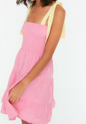 Femme portant une robe d'été rose à volants avec un corsage smocké et des bretelles jaunes nouées aux épaules, tenant l'ourlet de la robe d'une main.