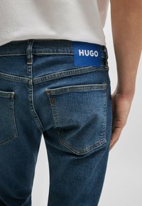 Tummansiniset farkut, joissa on sileä pinta. Takavyötäröllä on sininen logo-etiketti, jossa lukee "HUGO", ja standardit takataskut.