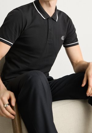 Polo noir avec bordures blanches, confectionné dans un tissu doux. Comprend un col classique et une patte à trois boutons, avec un logo sur la poitrine.