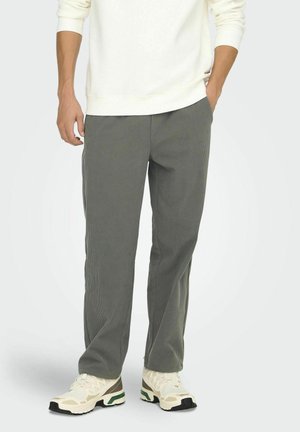 Homme portant un pantalon gris à fines rayures, des baskets blanches et une chemise blanche à manches longues, une main dans la poche, debout sur un fond uni.