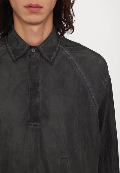 Chemise en tissu noir froissé avec un col boutonné, une patte de boutonnage cachée et un logo brodé dans le coin inférieur droit. Fini texturé.
