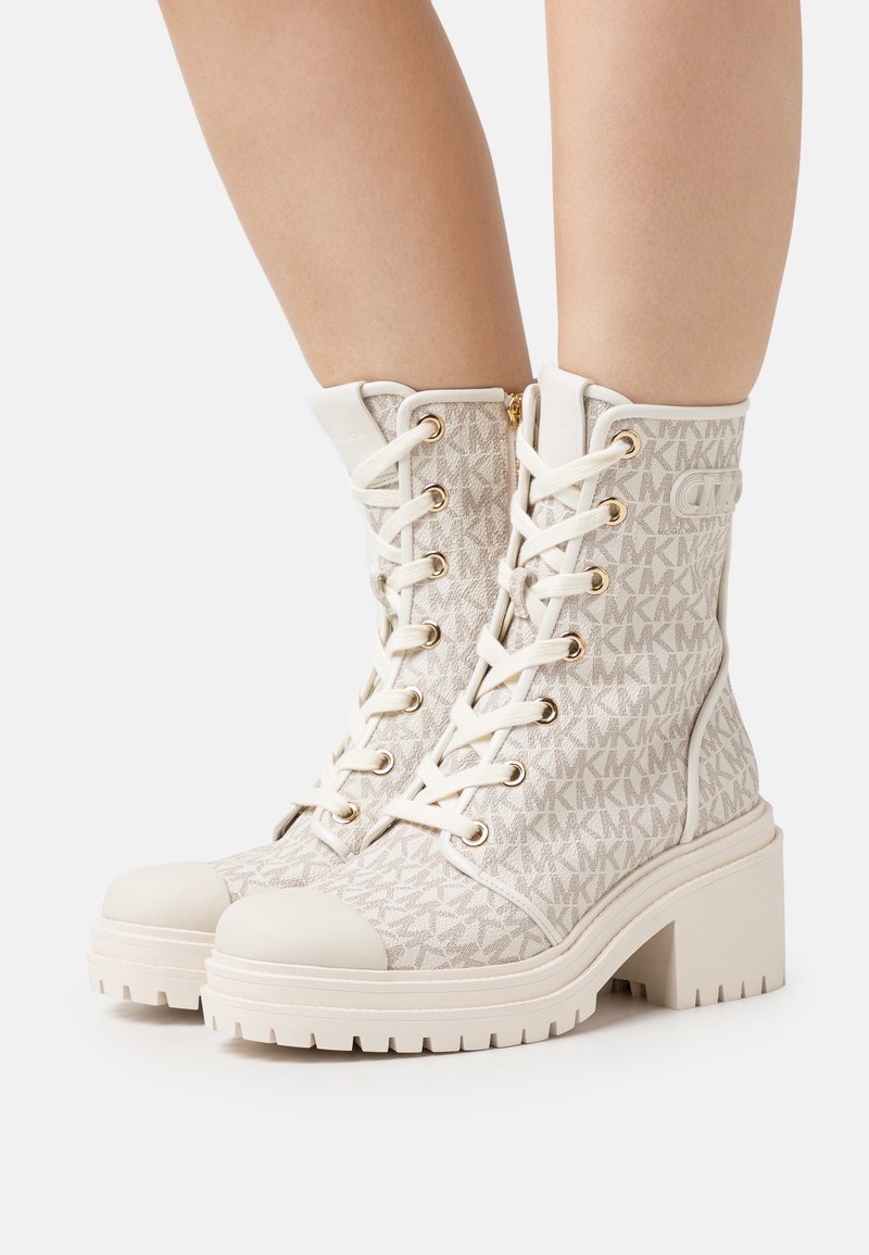 Bottes beiges montantes à lacets avec talons épais et motif logo MK portées sur jambes nues sur fond blanc.