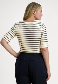 Sieviete ar blondu matu, valkājot krēmkrāsas un melnas svītrotas īsām piedurknēm krekliņu un tumšas augsta vidukļa bikses, skatās sāniski ar roku uz gurna.