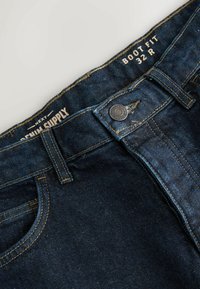 Jeans din denim albastru închis, cu croială pentru cizme, având cusături galbene, închidere cu nasture metalic și etichetă care indică mărimea 32 R.