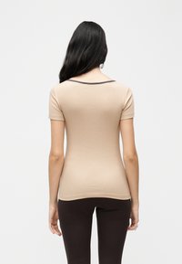 Γυναίκα με μακριά σκούρα μαλλιά, ντυμένη με κοντομάνικο μπεζ τοπ με μαύρο περίγραμμα και σκούρο καφέ παντελόνι, στέκεται με την πλάτη γυρισμένη σε απλό φόντο.