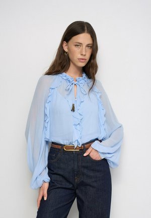 Mujer con cabello largo y castaño, que lleva una blusa ligera azul celeste con volantes y vaqueros oscuros con cinturón marrón, de pie frente a un fondo liso.