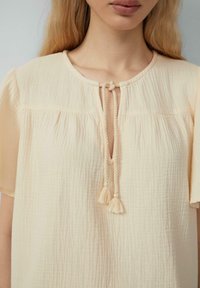 Blusa color crema, con texture, colletto rotondo, maniche corte e chiusura con cravatta con nappine sul davanti.
