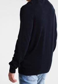 Pull en tricot bleu marine avec manches longues, poignets côtelés et ourlet inférieur. Texture lisse, encolure ronde classique et design minimaliste.