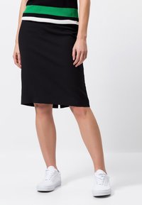 Schwarzer knielanger Rock aus weichem Stoff, mit einem seitlichen Schlitz für Bewegungsfreiheit, kombiniert mit weißen Sneakern für einen lässigen Look.