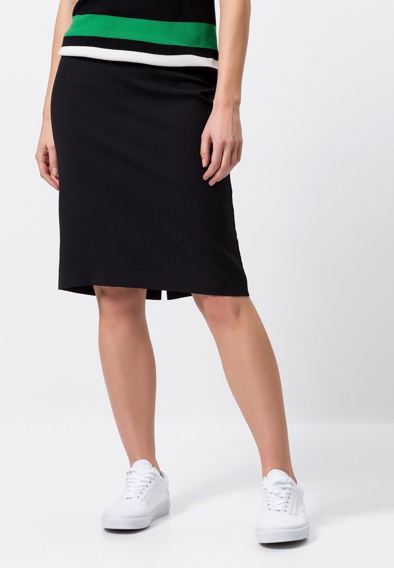 Schwarzer knielanger Rock aus weichem Stoff, mit einem seitlichen Schlitz für Bewegungsfreiheit, kombiniert mit weißen Sneakern für einen lässigen Look.