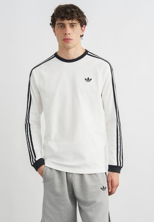 Junger Mann mit lockigem Haar trägt ein weißes langärmeliges Adidas-Shirt mit schwarzen Streifen und graue Adidas-Shorts, steht vor einem schlichten Hintergrund.