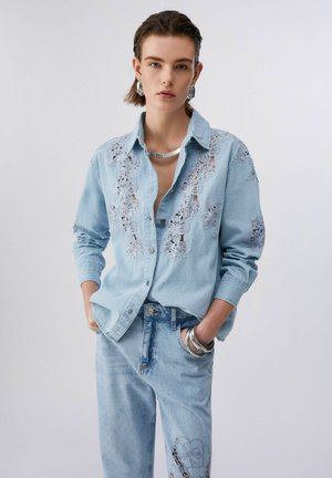 Junge Person trägt ein hellblaues, mit Spitze besticktes Denimhemd und Jeans, dazu Silberohrringe, eine Halskette und Armreifen, die Hände in den Taschen.