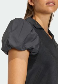 Czarny top z bufkami zrobionymi z teksturowanej tkaniny o subtelnym połysku i kołnierzykiem. Zbliżenie na ramię i rękę.