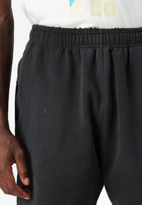 Črne bombažne trenirke z elastičnim pasom, stranskimi žepi in gladko teksturo. Imajo enojni šiv na tkanini.